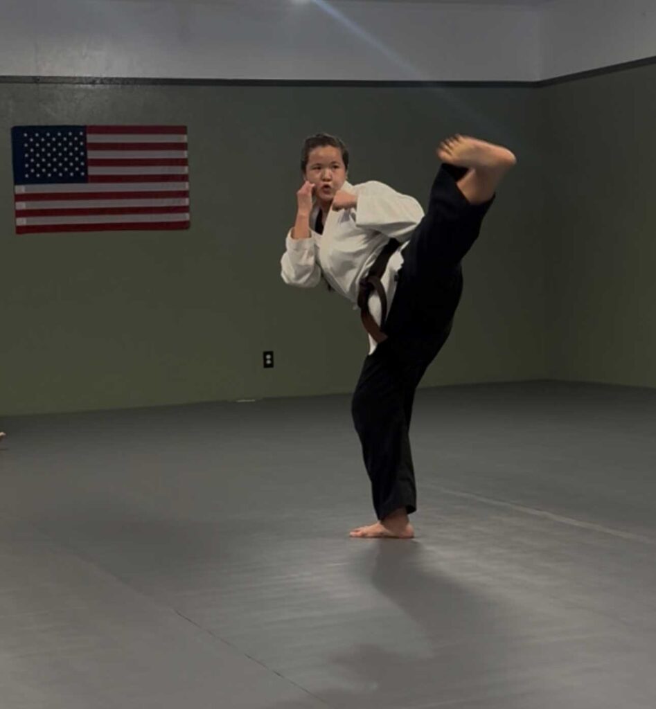 Woman practicing a karate kick in front of an American flag during Shin Nagare martial arts training.